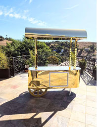 Glittering gold vendor cart with canopy and large decorative wheel on a sunlit patio terrace, ornate wrought-iron railing and rolling hillside views in the background