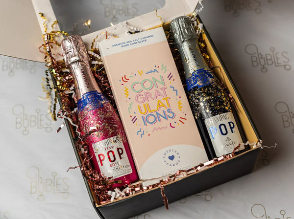Celebration gift box with two glitter-covered mini champagne bottles, a pastel "CONGRATULATIONS" chocolate bar and metallic confetti