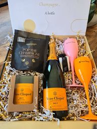 Luxury champagne gift box with orange-labeled bottle, pink and orange champagne flutes, 'Pop the Champagne' gourmet popcorn, boxed candle and gold-and-white shredded filler.