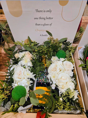 Festive champagne gift box with a glittery-neck bottle nestled in white roses, chrysanthemums and evergreen sprigs, with a sign that reads there is only one thing better than a glass of Champagne... A bottle.