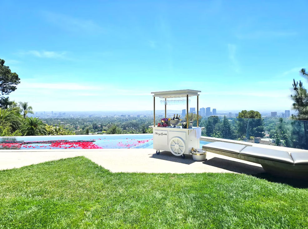 Hillside luxury infinity pool with rose petals, a white mobile drink cart and lounge chair overlooking a sprawling city skyline under a bright blue sky.