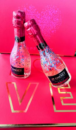Two mini sparkling-wine bottles in pink foil, sprinkled with blue and pink glitter, posed on a hot-pink gift box with gold lettering and a dotted heart — festive Valentine’s display.