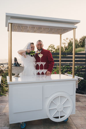 Bride in a veil pours champagne into a stacked coupe-glass tower as a smiling groom in a red jacket stands behind a white wedding cart with gold accents on an outdoor terrace at sunset
