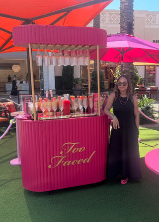 Person in sunglasses poses beside a hot-pink pop-up drink cart with hanging wine glasses, champagne flutes and bottles under pink and orange umbrellas in an outdoor shopping plaza