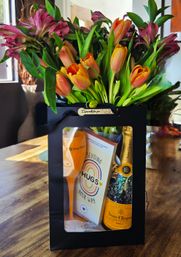 Bright orange tulips and magenta blooms in a black gift bag on a wooden dining table, with a sparkling wine bottle, orange flute, and a card reading Sending Hugs Your Way.