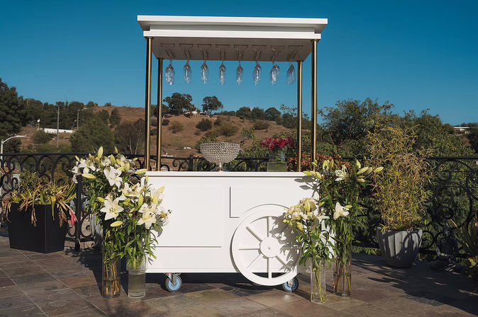 Chic white mobile drink cart with hanging wine glasses and crystal bowl, flanked by tall white lilies on a sunny outdoor terrace overlooking rolling hills.