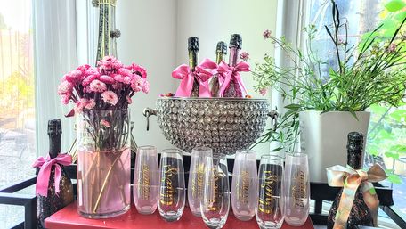 Sunlit indoor party setup with a crystal-studded champagne ice bucket holding three pink-ribboned sparkling bottles, a glass vase of pink chrysanthemums, potted greenery, and a row of 'Cheers' champagne flutes on a red cart.