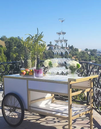 Sunny terrace cart set for celebration with a towering champagne coupe pyramid, ice bucket and bottle, pineapple and mangoes, white floral arrangements overlooking green hills
