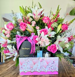 Vibrant pink-and-white bouquet of roses, lilies and snapdragons arranged in a decorative pink gift box on a wooden table, topped with a celebratory ribbon and a luxury champagne gift — festive centerpiece.
