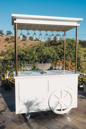White mobile outdoor bar cart with canopy and brass posts, hanging wine glasses and champagne bottles in a crystal ice bucket on a sunny patio overlooking a hillside