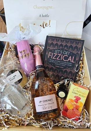Birthday bubbly gift box with rosé sparkling wine, 'Let's Get Fizzical' cocktail recipe book, pink 'Happy Birthday' flute, gummy bears and glassware nestled in gold-and-white crinkle paper.