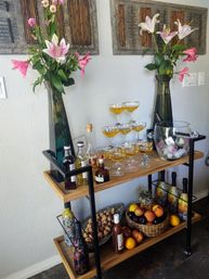 Wooden two-tier home bar cart in a dining nook with two tall vases of pink lilies, a champagne coupe tower of golden cocktails, assorted spirits and decorative bottles, an ice bucket with a chilled bottle, and a basket of fresh fruit.