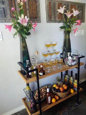 Wooden two-tier home bar cart in a dining nook with two tall vases of pink lilies, a champagne coupe tower of golden cocktails, assorted spirits and decorative bottles, an ice bucket with a chilled bottle, and a basket of fresh fruit.
