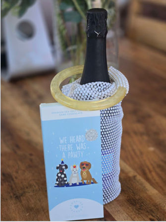 Sparkling champagne bottle in a rhinestone mesh sleeve with a gold bangle at the neck, paired with a blue dog-themed "We heard there was a pawty" greeting card on a wooden table.