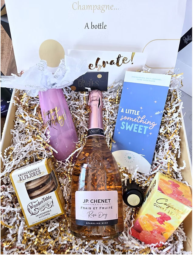 Birthday celebration gift box with a rosé sparkling wine bottle, pink "Happy Birthday" tumbler, chocolate bar, cookies and gummy candies nestled in gold-and-white shredded paper