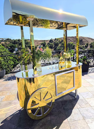 Gold mirrored beverage cart with canopy on a sunny outdoor terrace patio, decorated with floral arrangements and wine glasses, reflecting ornate railing and rolling green hills in the background.