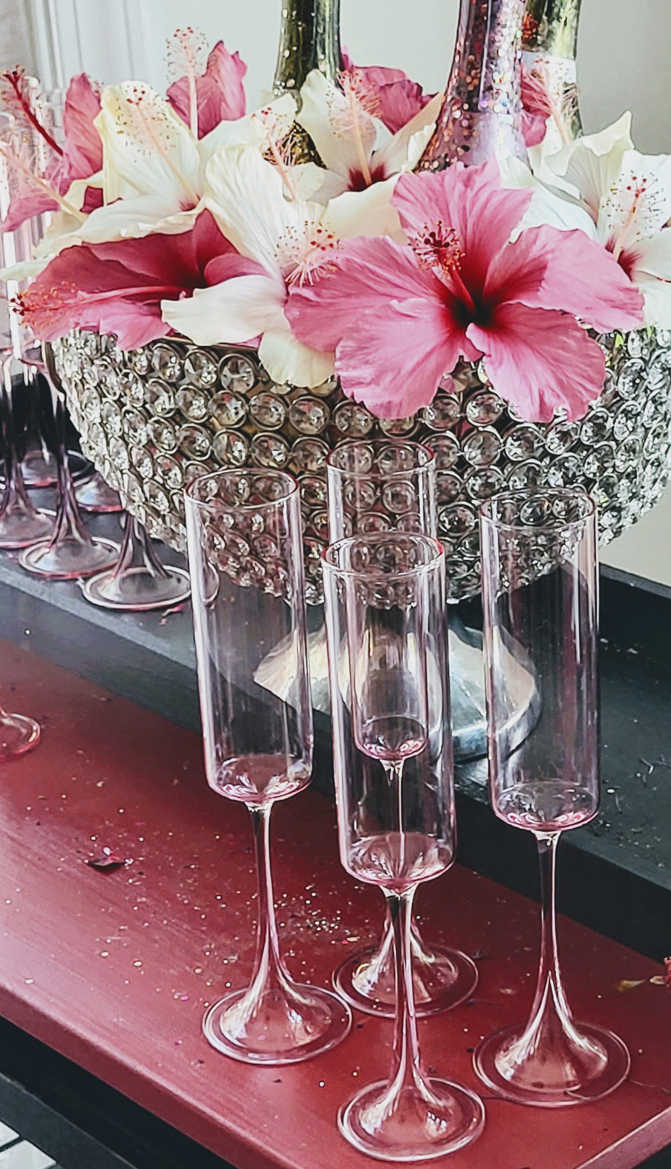 Sparkling crystal bowl centerpiece of pink and white hibiscus with translucent pink champagne flutes on a red table, festive party decor.