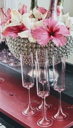 Sparkling crystal bowl centerpiece of pink and white hibiscus with translucent pink champagne flutes on a red table, festive party decor.