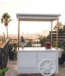White mobile rooftop bar cart with gold canopy, crystal ice bucket holding champagne, stacked martini glasses and a red flower box on a sunset terrace overlooking a distant city skyline and palm tree