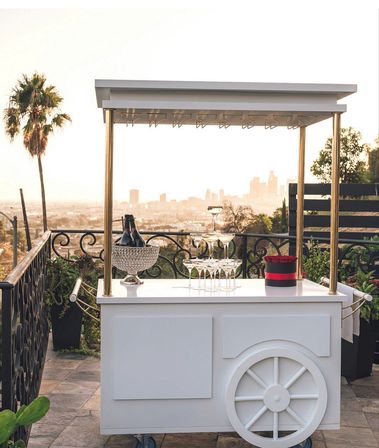 White mobile rooftop bar cart with gold canopy, crystal ice bucket holding champagne, stacked martini glasses and a red flower box on a sunset terrace overlooking a distant city skyline and palm tree