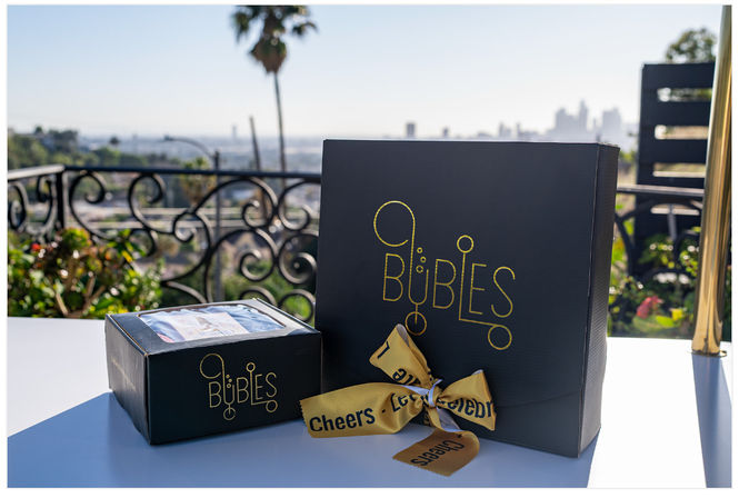 Two black gift boxes with gold lettering and a yellow "Cheers" ribbon on a sunlit balcony table, ornate wrought-iron railing, palm tree and distant city skyline in the background.