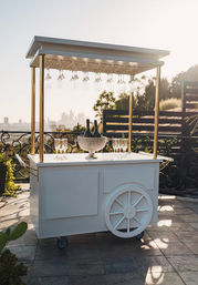 Chic white mobile rooftop bar cart with hanging wine glasses and crystal ice bucket holding champagne, set on a terrace overlooking a city skyline at sunset.