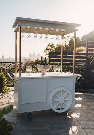 Chic white mobile rooftop bar cart with hanging wine glasses and crystal ice bucket holding champagne, set on a terrace overlooking a city skyline at sunset.