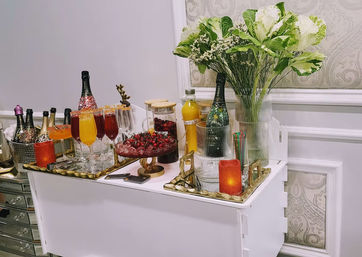 Stylish indoor drink station on a white console table with champagne bottles in ice, colorful cocktail flutes, a bowl of fresh berries, orange juice pitcher, lit candle and a tall green-and-white floral arrangement.