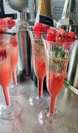 Three bubbly pink raspberry champagne cocktails in tall flutes garnished with fresh raspberries and mint on a metallic bar counter.