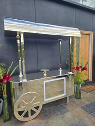 Gleaming stainless steel cart with curved canopy and decorative wheel on a stone patio by a dark exterior wall and wooden door, flanked by tall vases of red flowers and bamboo - modern outdoor serving setup