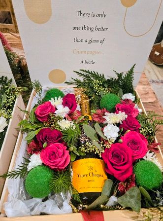 Wooden gift box with a champagne bottle nestled among bright pink roses, white filler flowers, green moss balls and festive greenery; decorative lid shows a champagne quote.