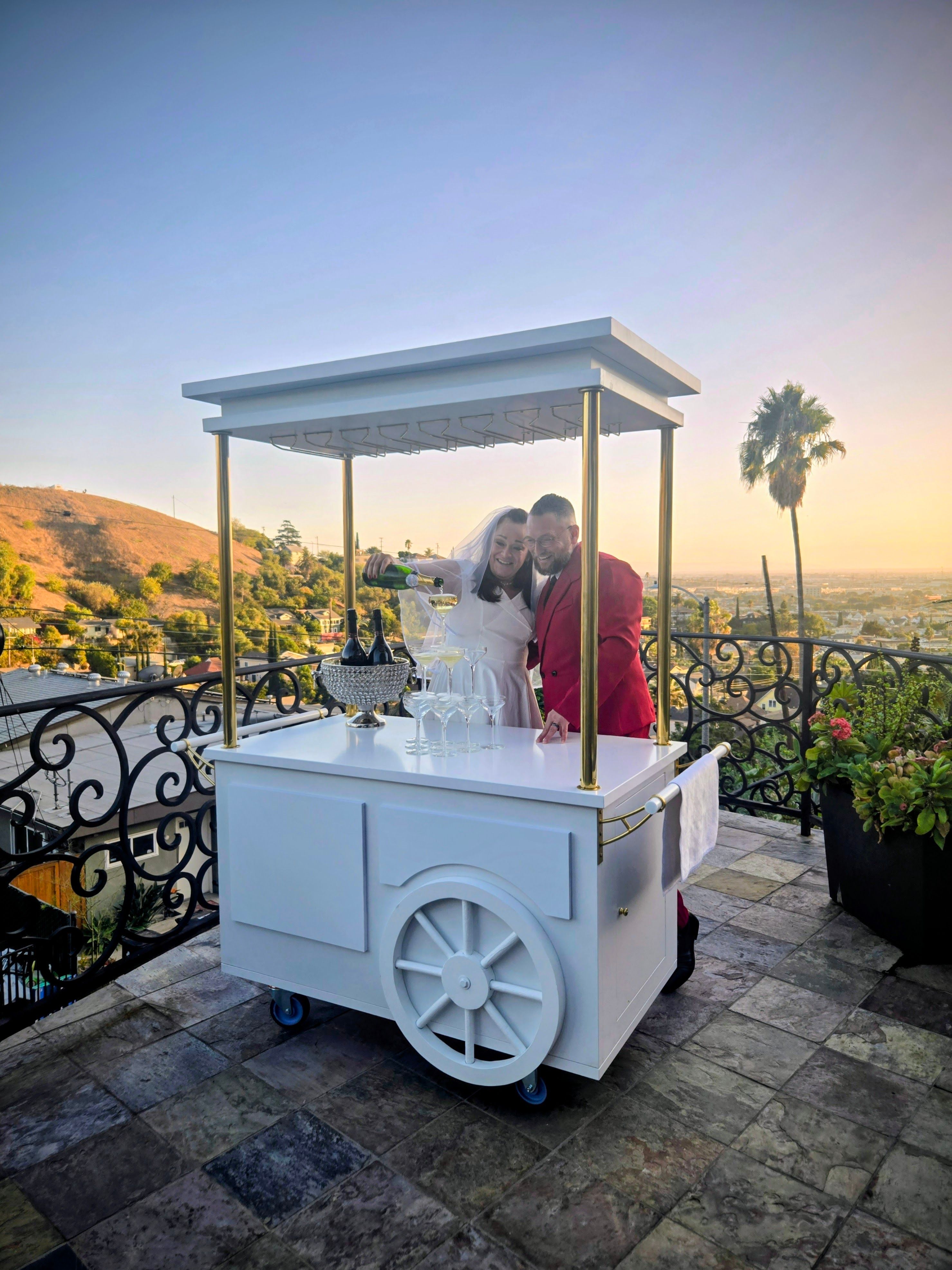 Couple in wedding attire pouring champagne from a bottle into a stack of glasses on a white mobile cart at a rooftop terrace at sunset, overlooking rolling hills, a palm tree and city skyline.