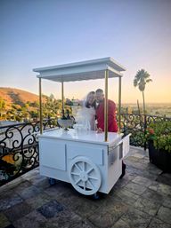 Couple in wedding attire pouring champagne from a bottle into a stack of glasses on a white mobile cart at a rooftop terrace at sunset, overlooking rolling hills, a palm tree and city skyline.