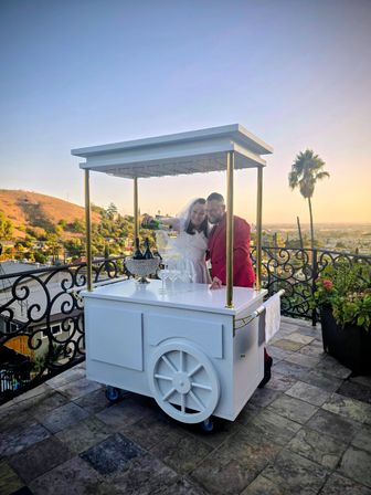 Couple in wedding attire pouring champagne from a bottle into a stack of glasses on a white mobile cart at a rooftop terrace at sunset, overlooking rolling hills, a palm tree and city skyline.