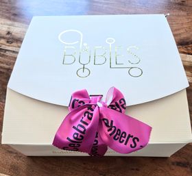White gift box with gold foil bubble graphic and bright pink satin ribbon tied in a bow with black celebratory lettering, sitting on a wooden table — stylish party gift box.