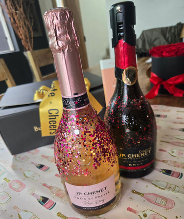 Two sparkling wine bottles on a celebratory table — a pink rosé bottle coated in pink glitter next to a dark brut bottle, surrounded by gift boxes and a red ribboned rose box.