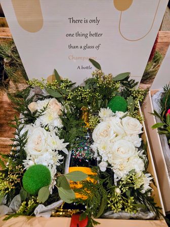 Gift box with a sparkling wine bottle topped in glitter, nestled among white roses, chrysanthemums and evergreen greenery, accented with green pom-poms and a decorative sign with a playful champagne quote.
