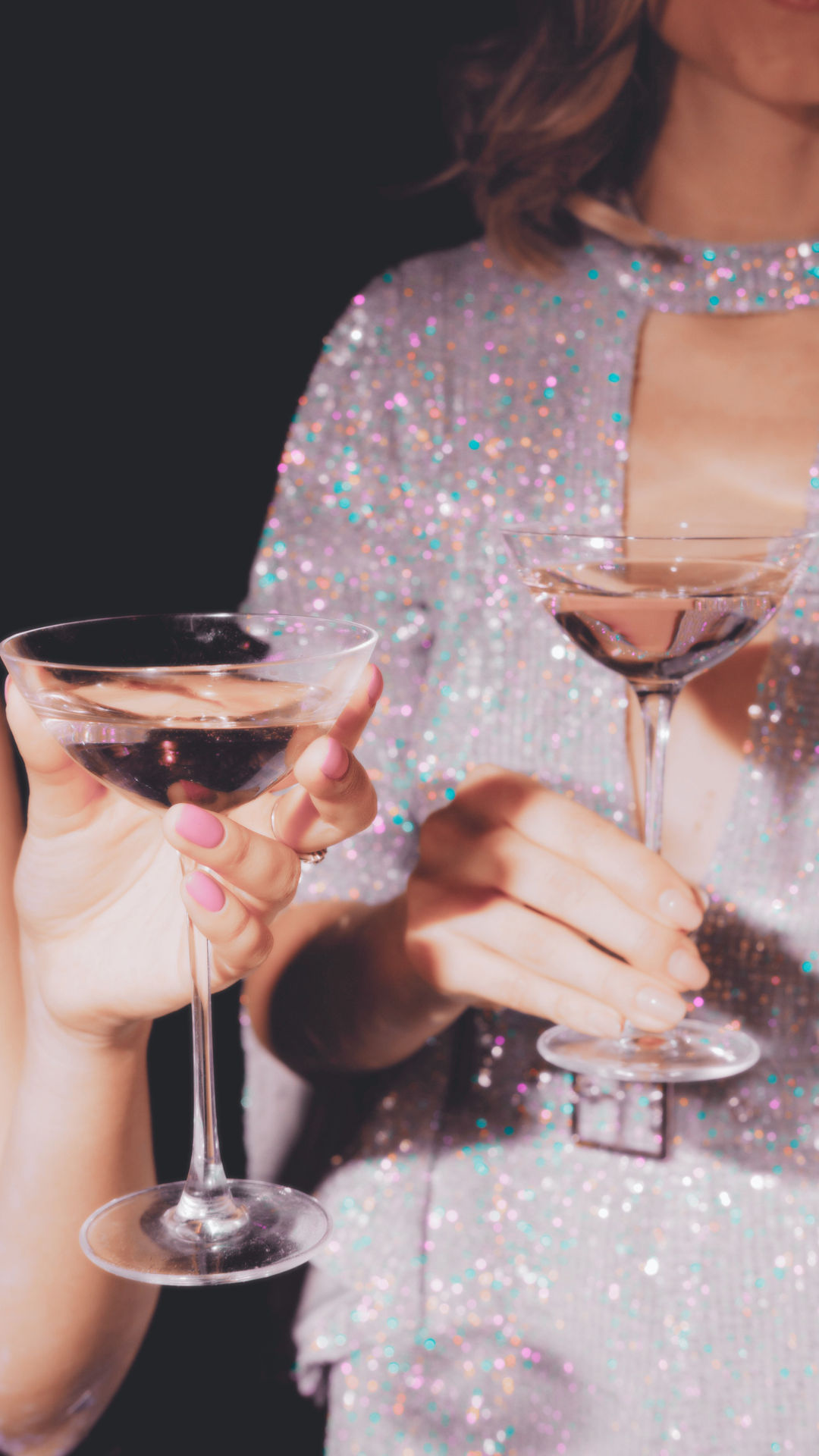 Close-up of two hands with pink manicures holding coupe cocktail glasses of champagne against a glittering sequined dress, glamorous night-out celebration.