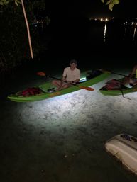Night kayaking: paddler in a glowing green kayak with orange paddle in shallow clear coastal water near mangroves, distant city lights reflecting.