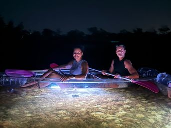 Two kayakers at night in a transparent kayak lit from below, smiling and paddling with pink paddles over illuminated shallow water