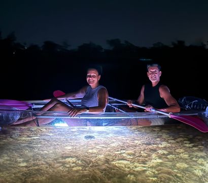Two kayakers at night in a transparent kayak lit from below, smiling and paddling with pink paddles over illuminated shallow water