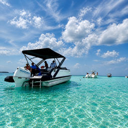 Sun-soaked motorboat with canopy anchored in shallow, crystal-clear turquoise water as people relax under a bright blue sky dotted with fluffy white clouds — tropical beach getaway vibe.