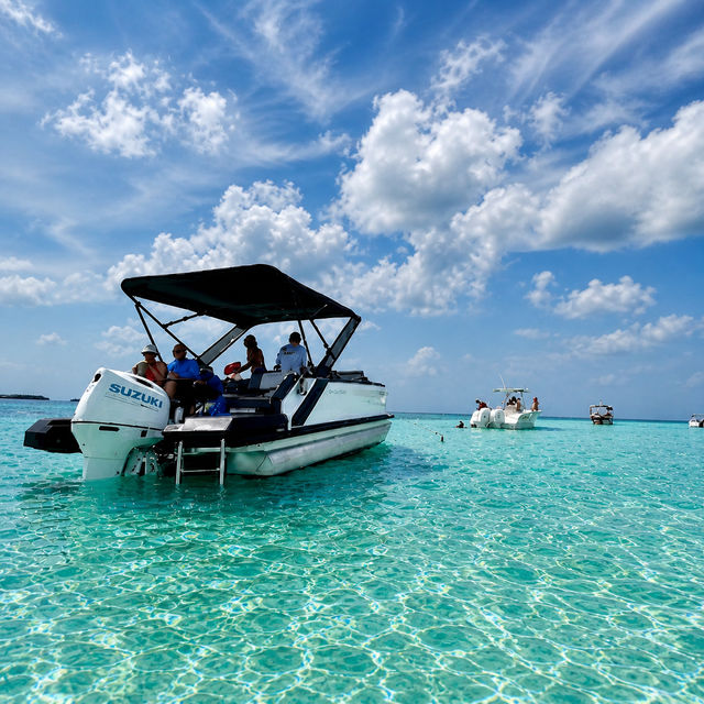 Sun-soaked motorboat with canopy anchored in shallow, crystal-clear turquoise water as people relax under a bright blue sky dotted with fluffy white clouds — tropical beach getaway vibe.