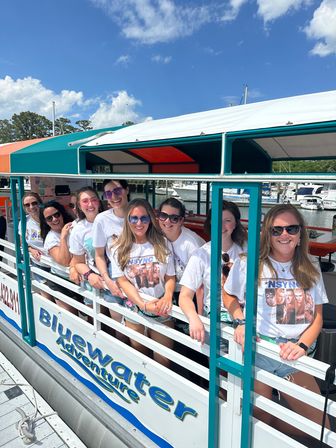 Smiling group of women in matching white graphic T‑shirts leaning on the railing of a colorful covered tour boat at a sunny marina, with yachts and blue sky in the background.
