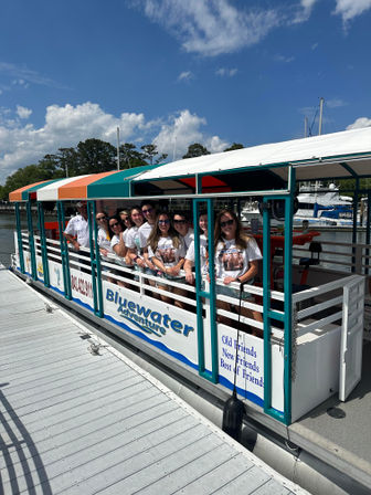 Group of friends in matching white shirts smiling aboard a colorful covered sightseeing boat docked at a sunny coastal marina with sailboats and blue sky