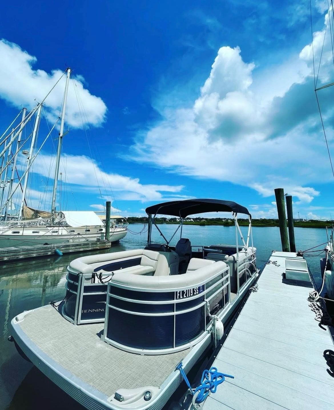 Pontoon boat docked at a sunny coastal marina beside a wooden pier, calm water reflecting sailboats under a bright blue sky with fluffy white clouds.