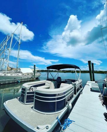 Pontoon boat docked at a sunny coastal marina beside a wooden pier, calm water reflecting sailboats under a bright blue sky with fluffy white clouds.
