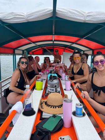 Group of women in swimsuits laughing around a central bar on a covered party boat with a red-and-teal canopy, drinks, a straw sun hat and phones on the counter as the boat cruises on calm water.