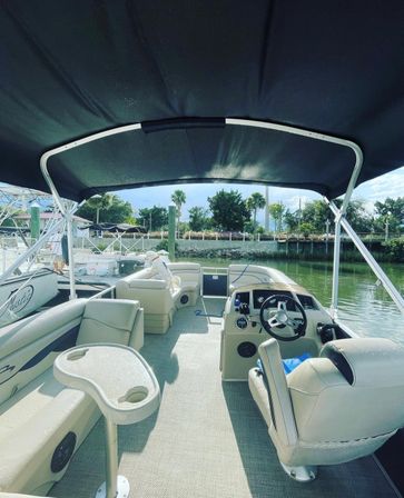 Cozy beige pontoon boat interior under a black bimini top, showing captain’s helm with steering wheel and swivel chair, molded cupholder table, bench lounges with a small white dog, docked at a sunny marina with palm trees and calm waterfront