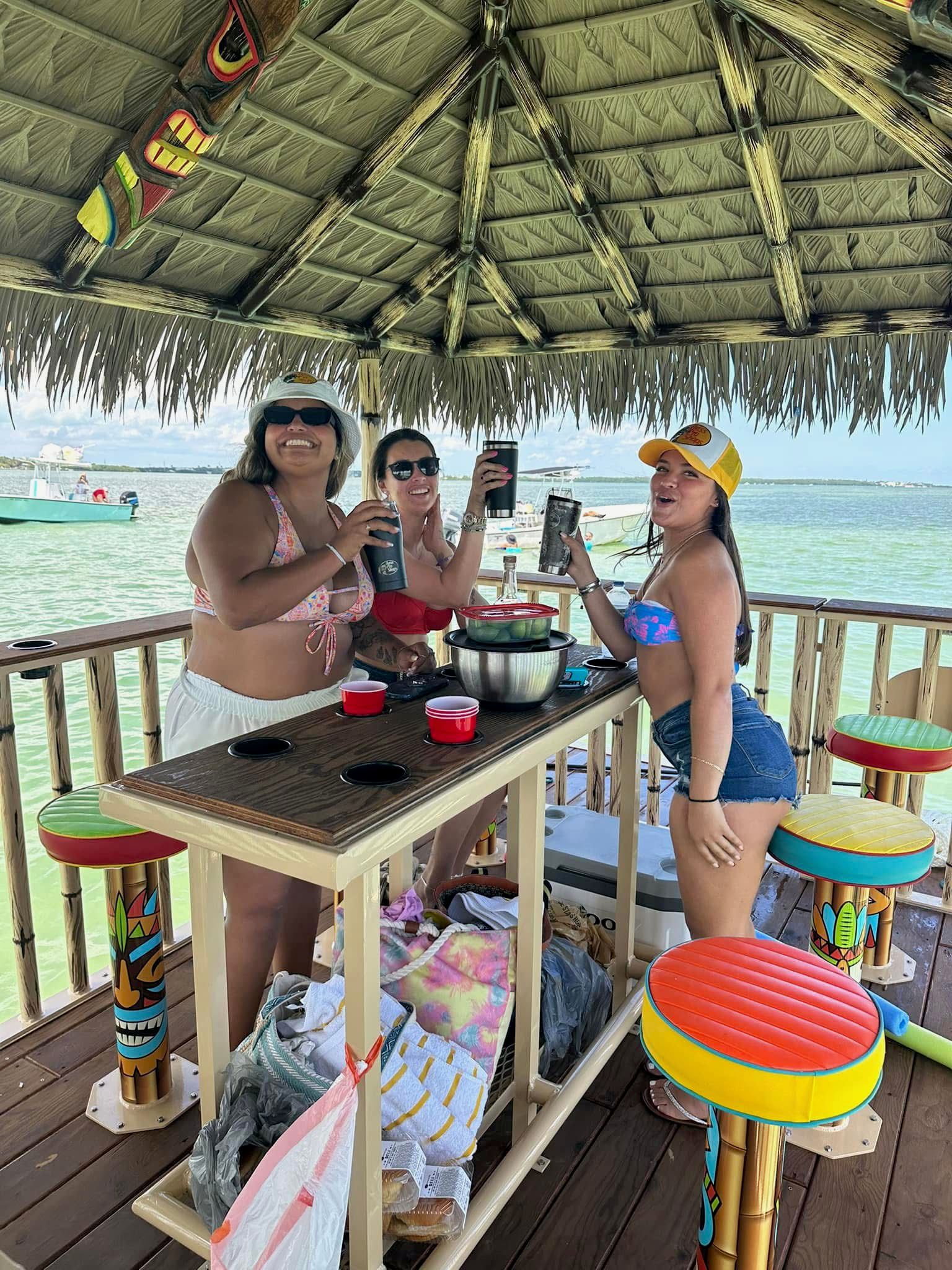 Three people in swimwear toasting drinks under a thatched tiki-hut bar above turquoise water, colorful stools and a boat in the bay.